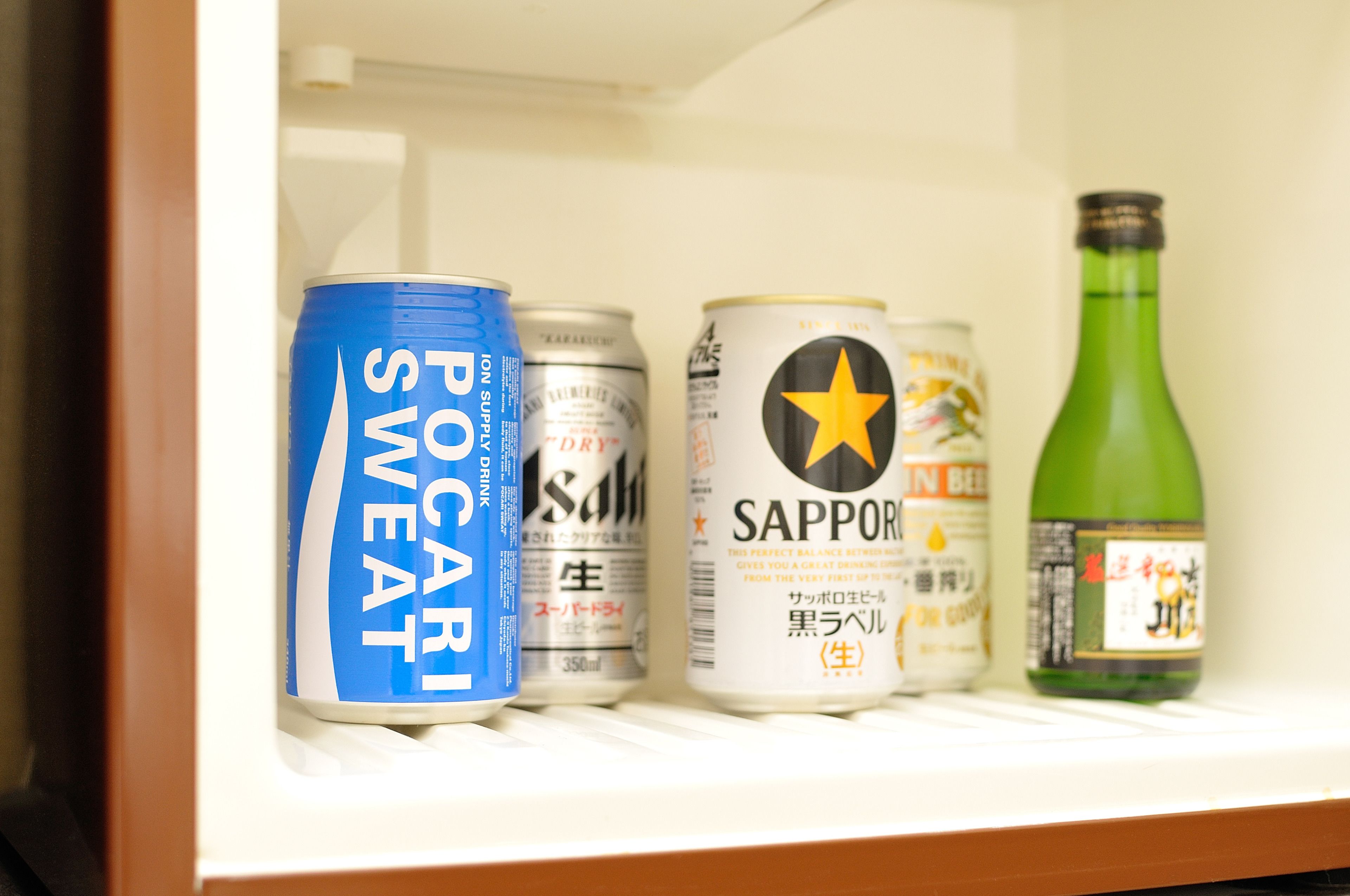 Estante refrigerador con bebidas, incluyendo una lata azul etiquetada “POCAR SWEAT”, una lata blanca etiquetada “SAPPORO” y una botella verde.