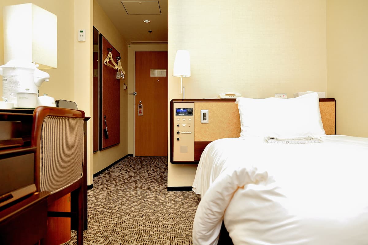 Ein Hotelzimmer mit Bett, Schreibtisch, Stuhl, Lampe, Nachttisch und einer an der Wand befestigten Steuerung.