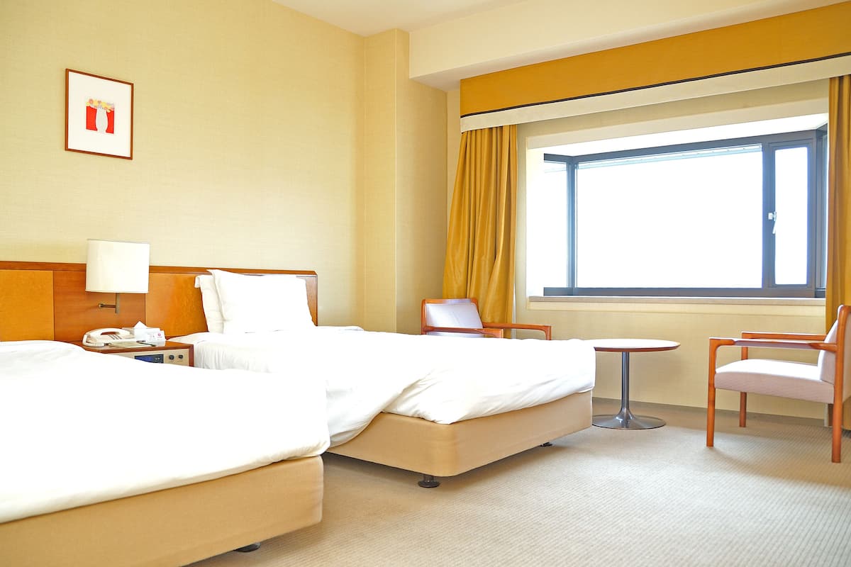 Ein Hotelzimmer mit zwei Betten, einem kleinen Tisch, einem Stuhl, einer Lampe und einem Fenster mit Vorhängen.