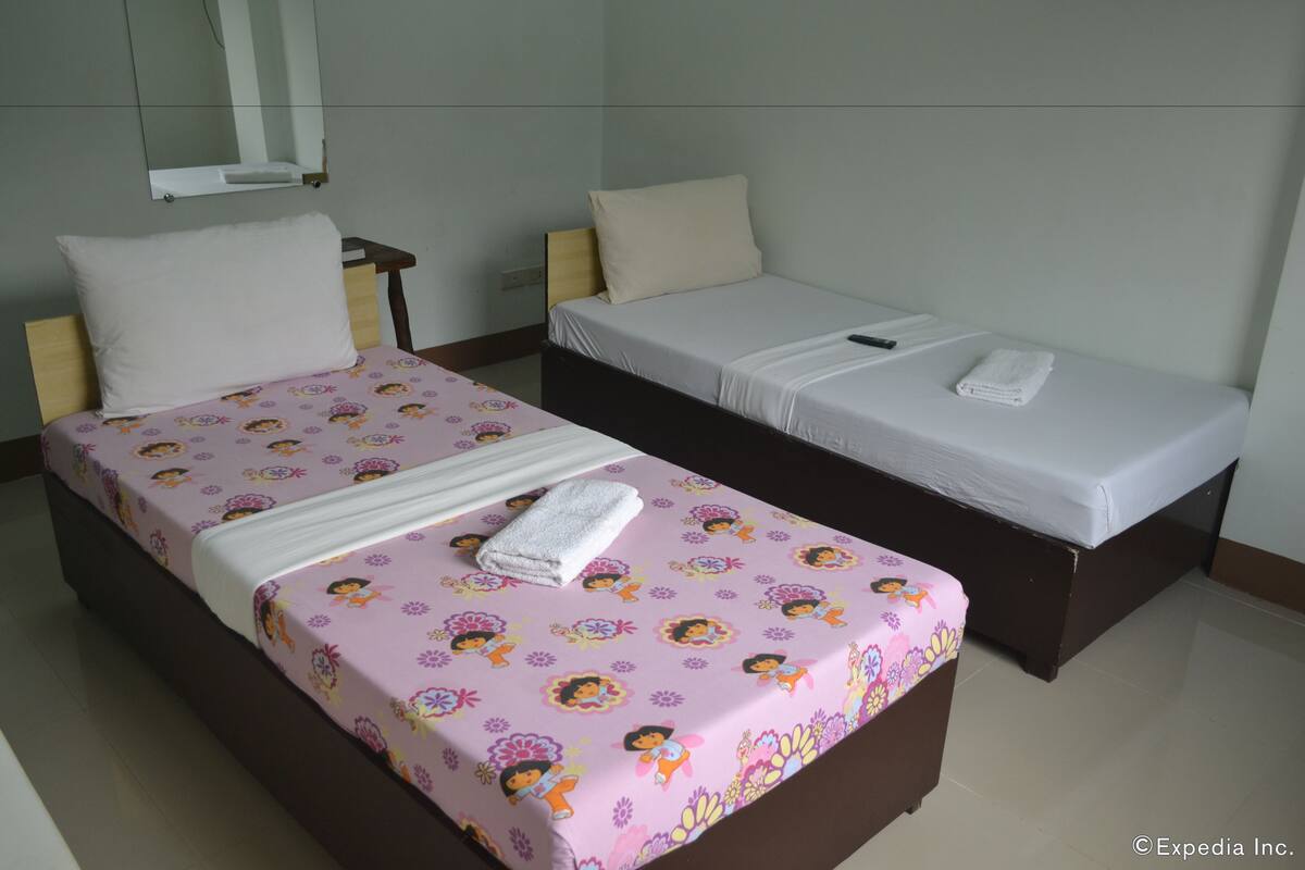 Ein Hotelzimmer mit zwei Betten, eines mit einer rosa floralen Tagesdecke und das andere mit einer weißen. Auf dem Bett mit der weißen Decke liegt ein weißes Handtuch.