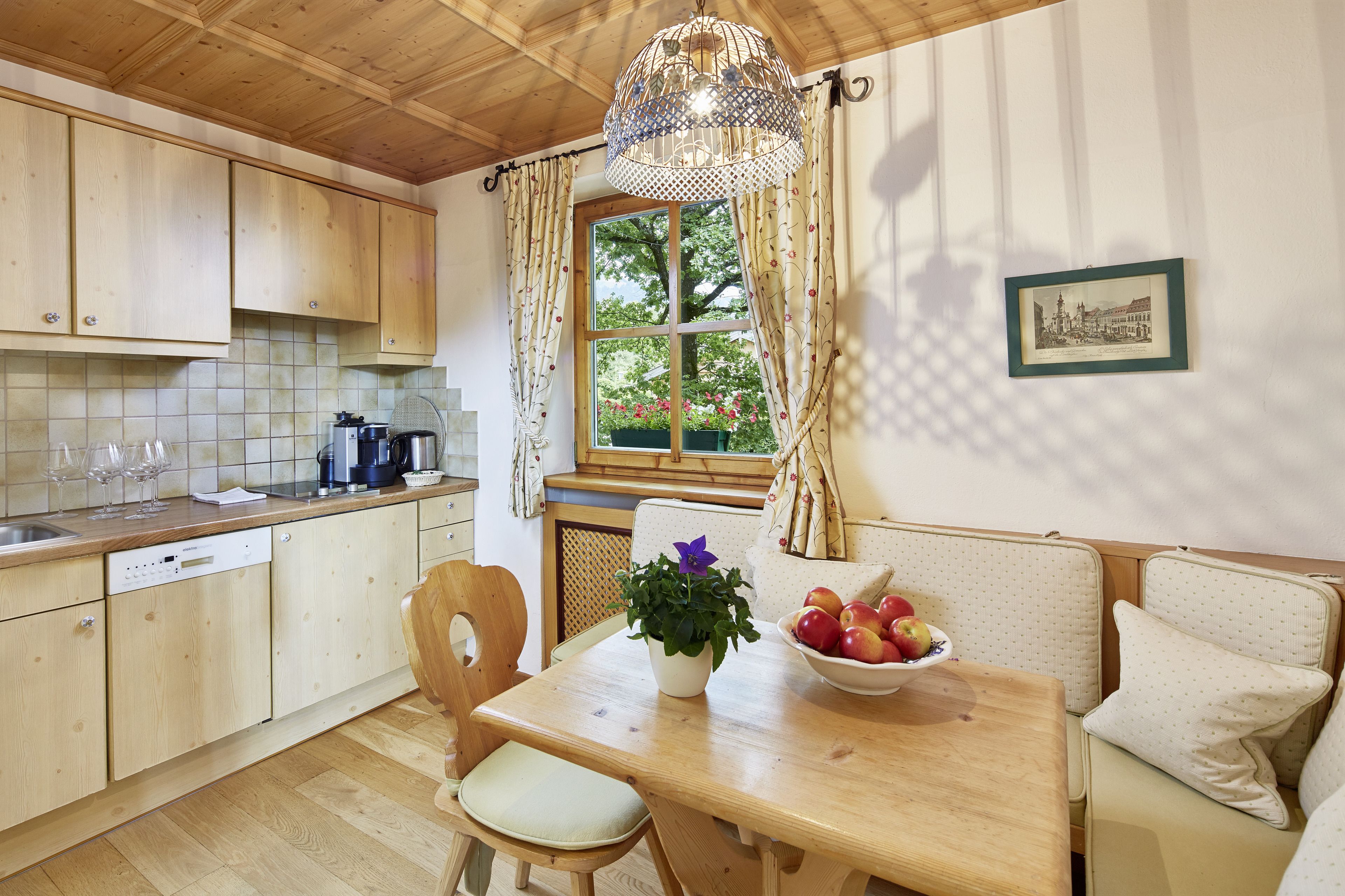 A kitchen with wooden cabinets, a dining table with a bowl of fruit, and a window with curtains.