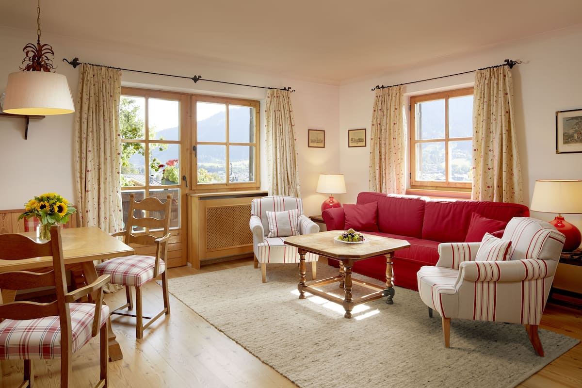 A cozy living room with a wooden dining table, a red sofa, striped armchairs, and a view of the outdoors through large windows.