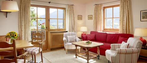 A cozy living room with a wooden dining table, a red sofa, striped armchairs, and a view of the outdoors through large windows.