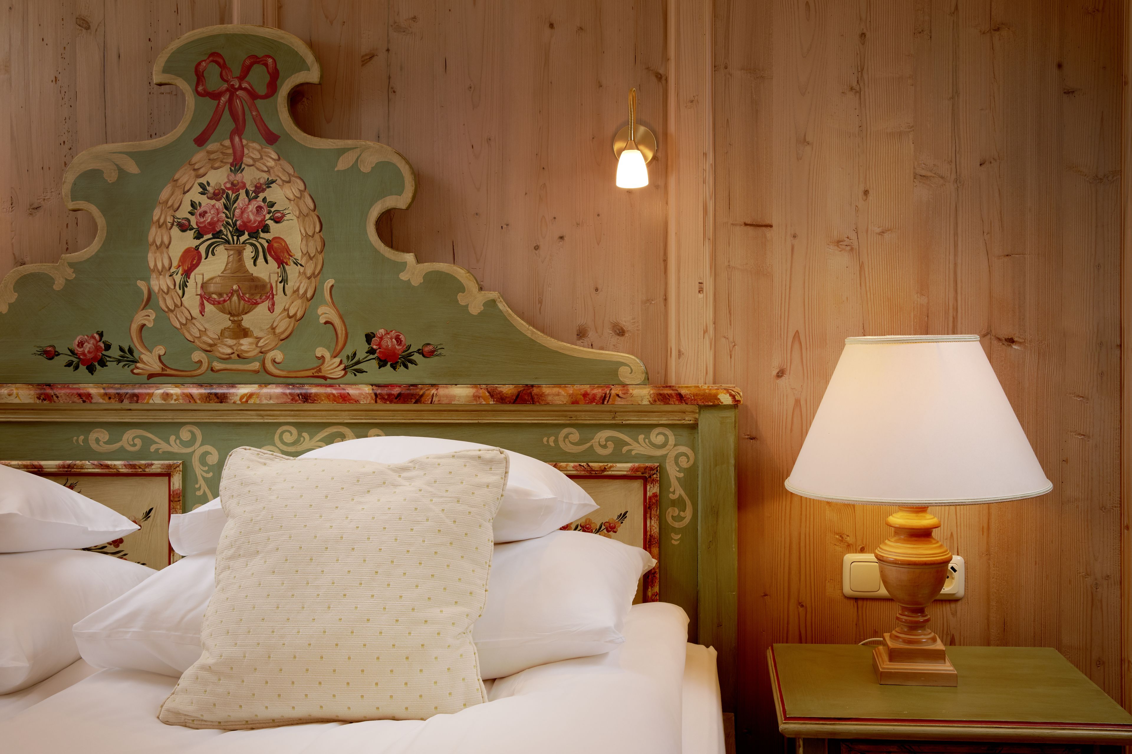 A neatly made bed with white linens and a single patterned pillow, a wooden nightstand with a lamp, and a decorative headboard with floral and ribbon designs.