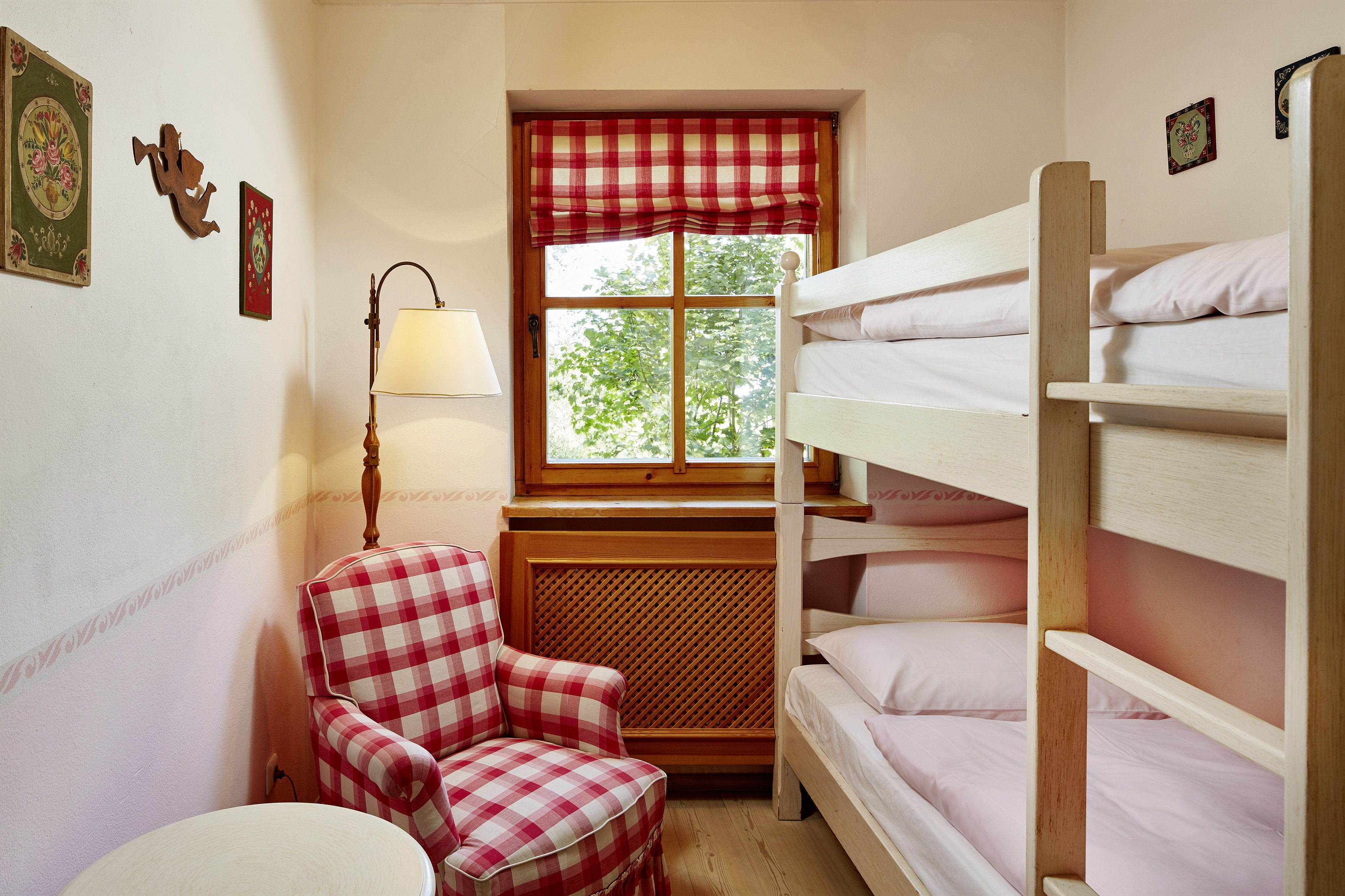 Une petite pièce agréable et chaleureuse, avec un lit superposé, une chaise, une table et une fenêtre agrémentée d’une lambrequin à carreaux.