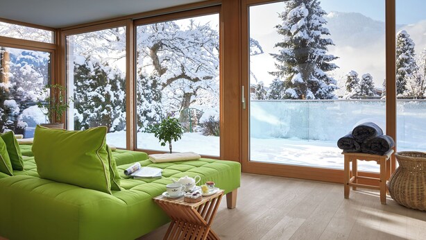 A modern living room with a green sofa, a small table with tea set, and a view of a snowy landscape through large windows. - Landsitz Römerhof - Hotel Apartments (Kitzbuhel)