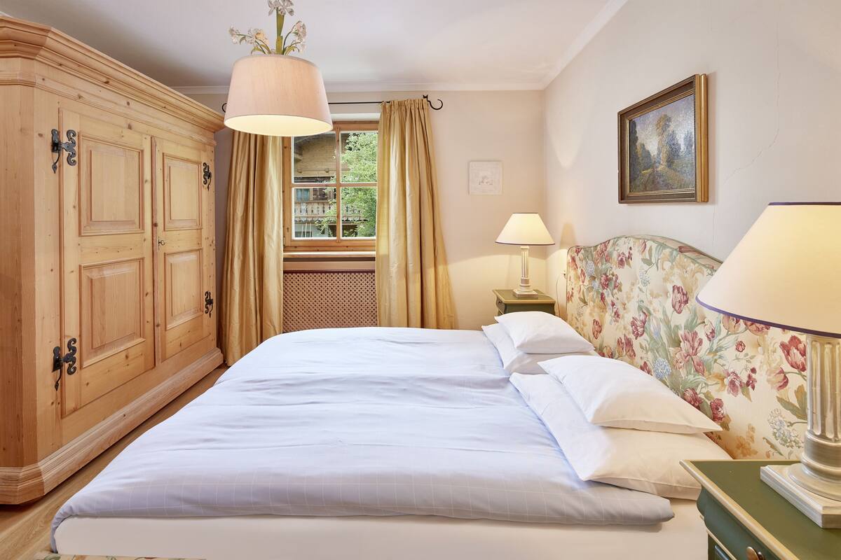 A bedroom with a large bed, wooden wardrobe, floral headboard, and a window with curtains.
