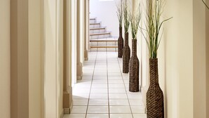 A long, narrow hallway with white walls and columns, leading to a staircase. There are several large, woven vases with tall grasses placed along the walls. - Landsitz Römerhof - Hotel Apartments (Kitzbuhel)