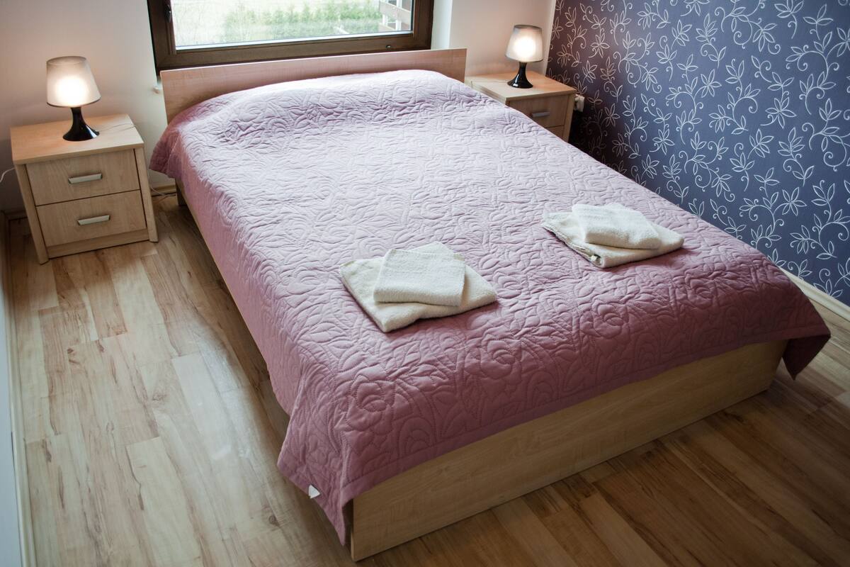 A neatly made bed with a pink quilt and two folded towels, wooden nightstands with lamps, and a window with a view of greenery.