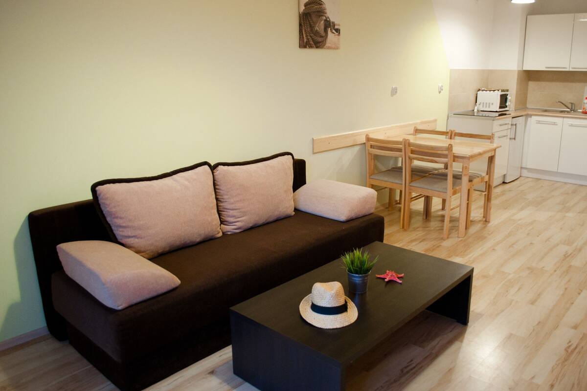 A living room with a dark brown sofa, two light brown cushions, a wooden dining table with four chairs, a small potted plant, and a decorative hat on a black coffee table.