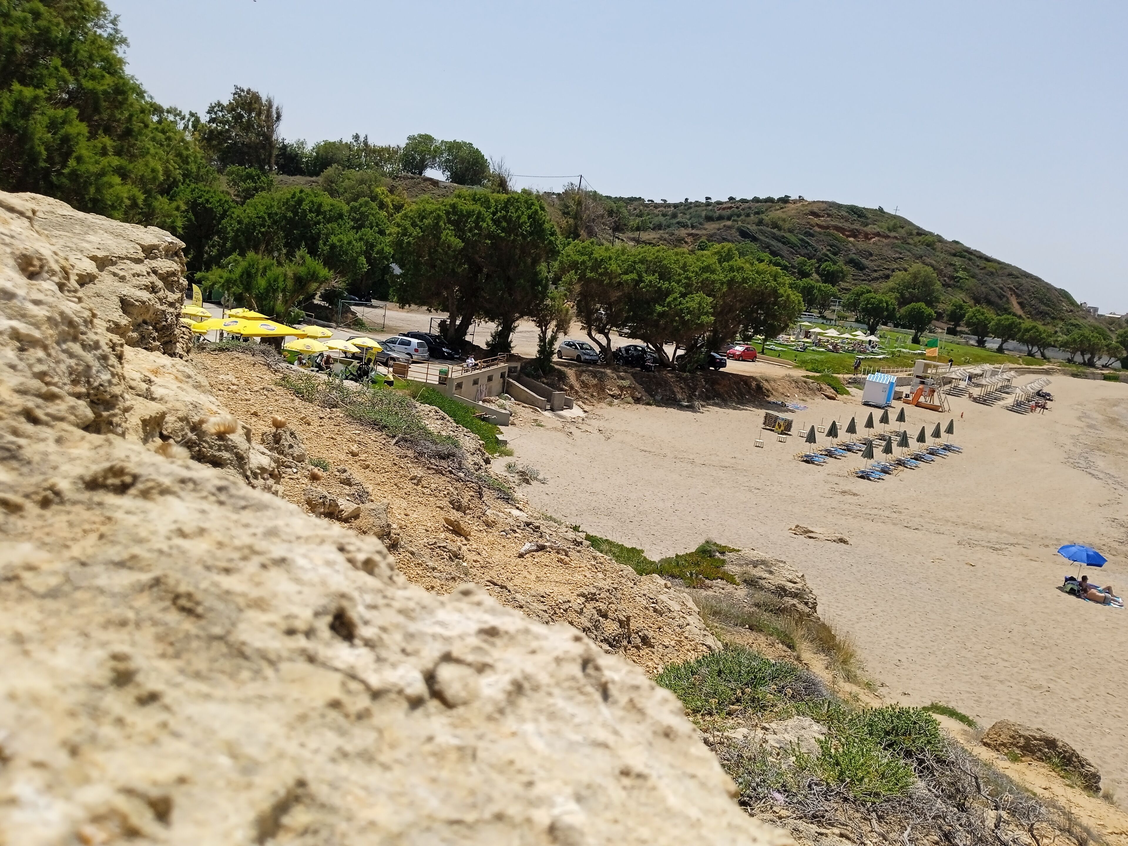 Une plage avec des parasols, des voitures et un premier plan rocheux.