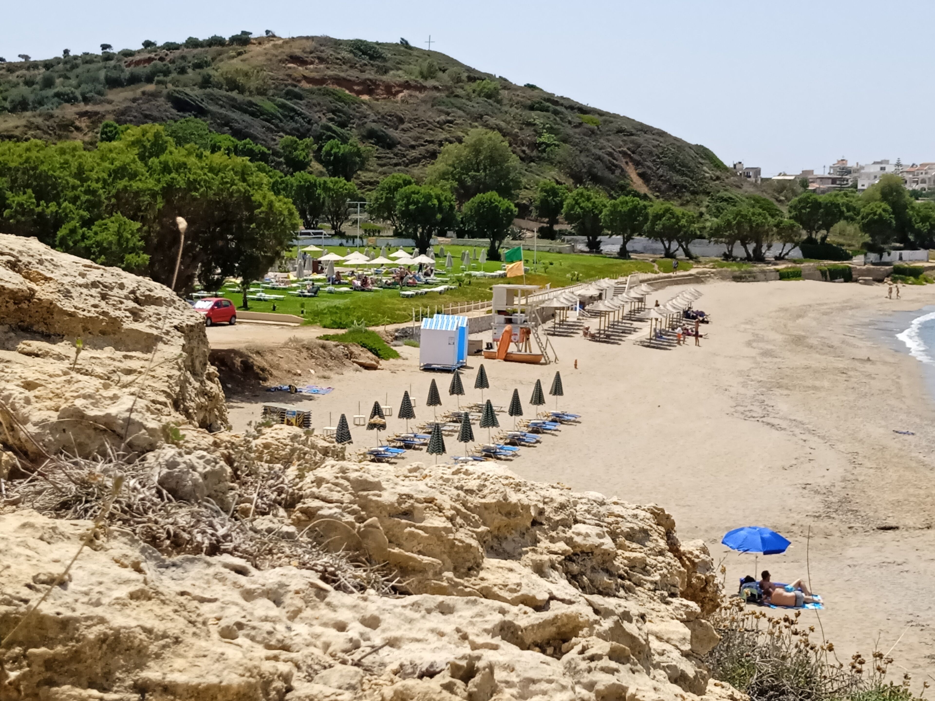 Ein Strand mit Reihen von Sonnenschirmen und Liegestühlen, einem Rettungsturm und einer sanften Hügelkette im Hintergrund.