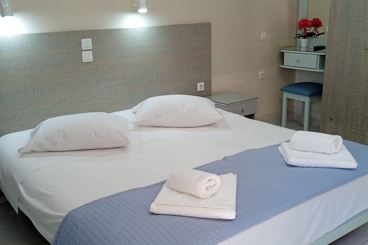A hotel room with a bed, two pillows, a folded towel, a nightstand, a mirror, and a potted plant.