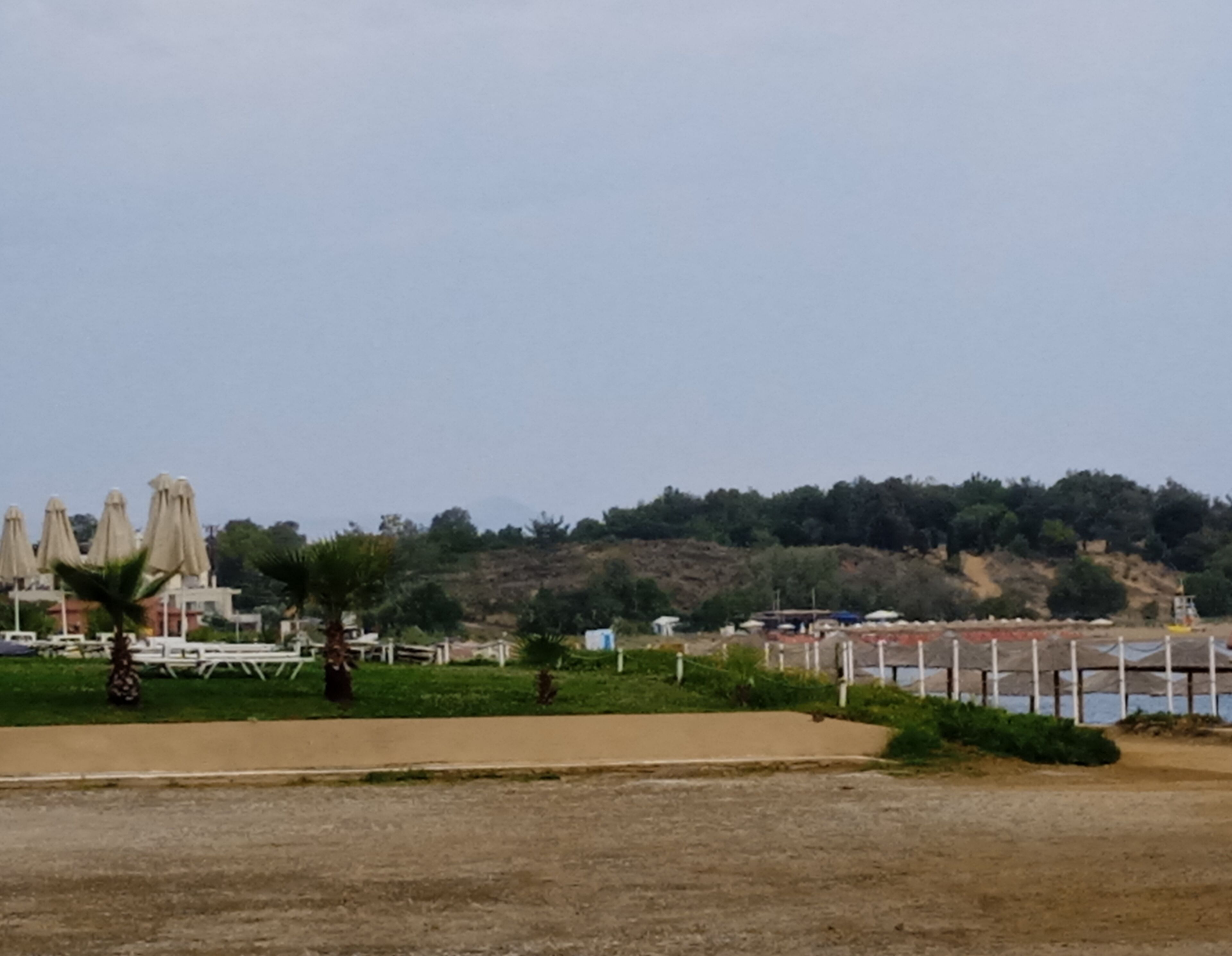 A beachside area with umbrellas and tables, a hill with trees, and a body of water in the background.