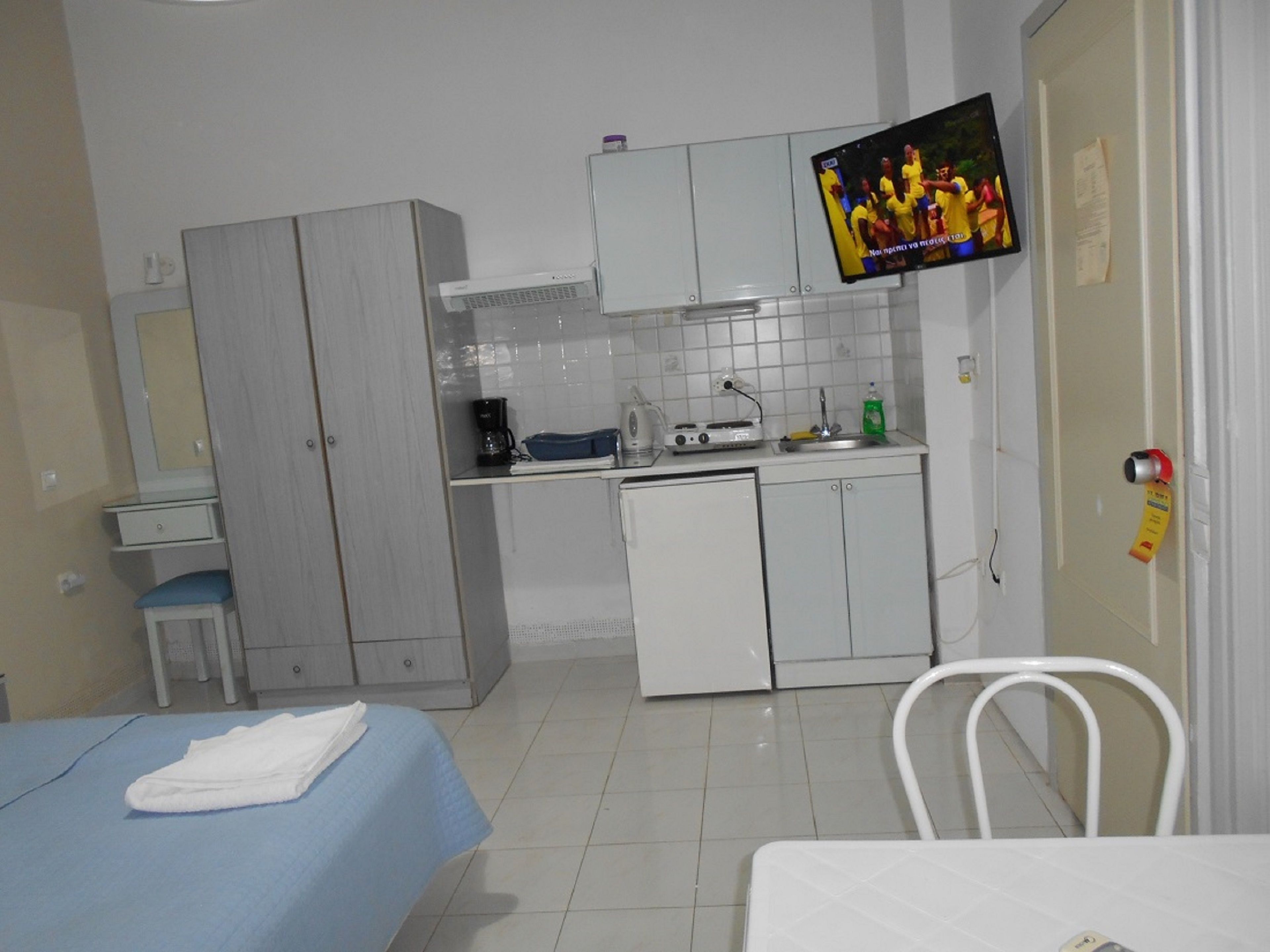 A compact hotel room with a kitchenette, a bed with a blue bedspread, a white chair, and a television mounted on the wall.