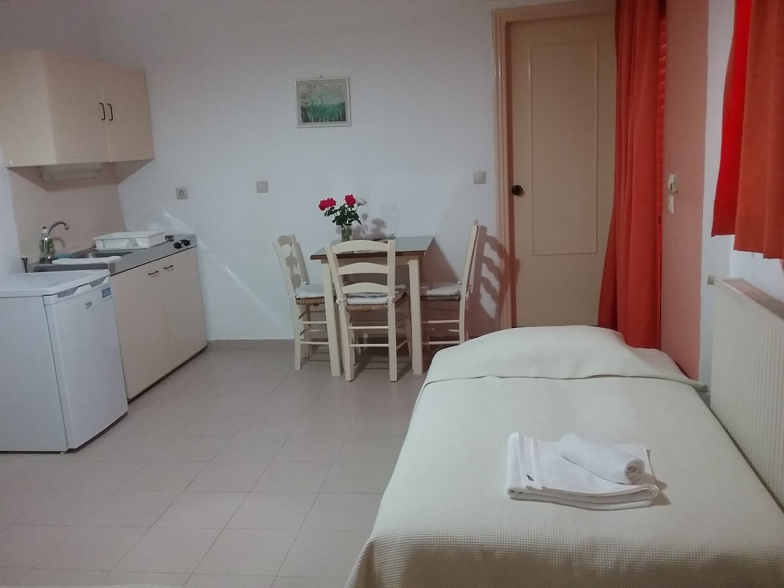 Una habitación pequeña y limpia con cama, escritorio y silla, kitchenette y una puerta con cortinas rojas.
