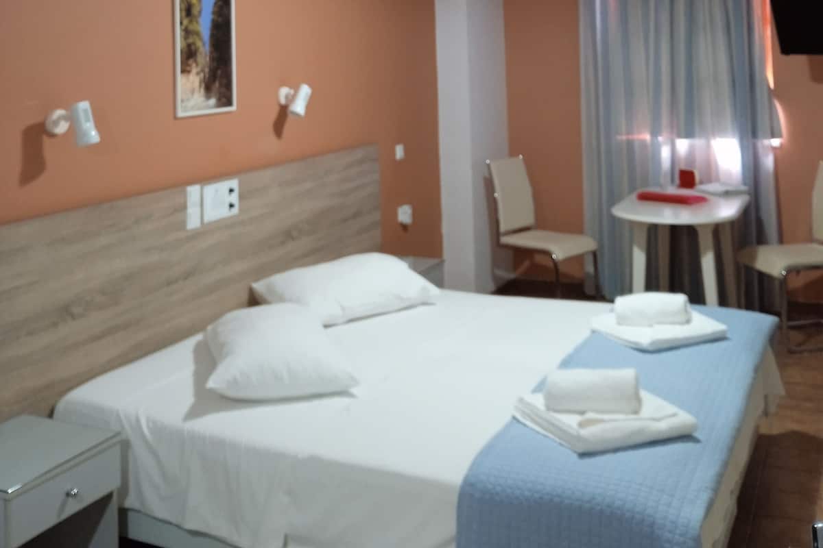 A hotel room with a bed, a small table, a chair, and a TV mounted on the wall.
