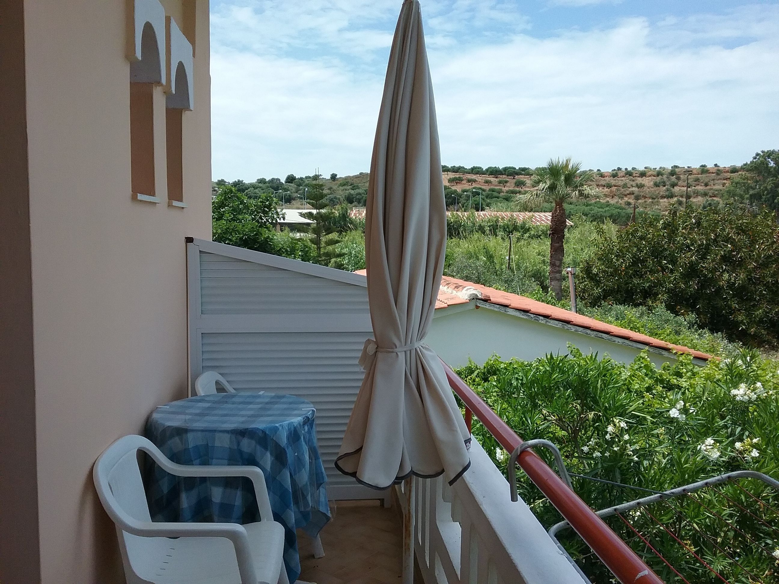 A balcony with a table and chairs, a beige umbrella, and a view of a green landscape with trees and a building in the distance.