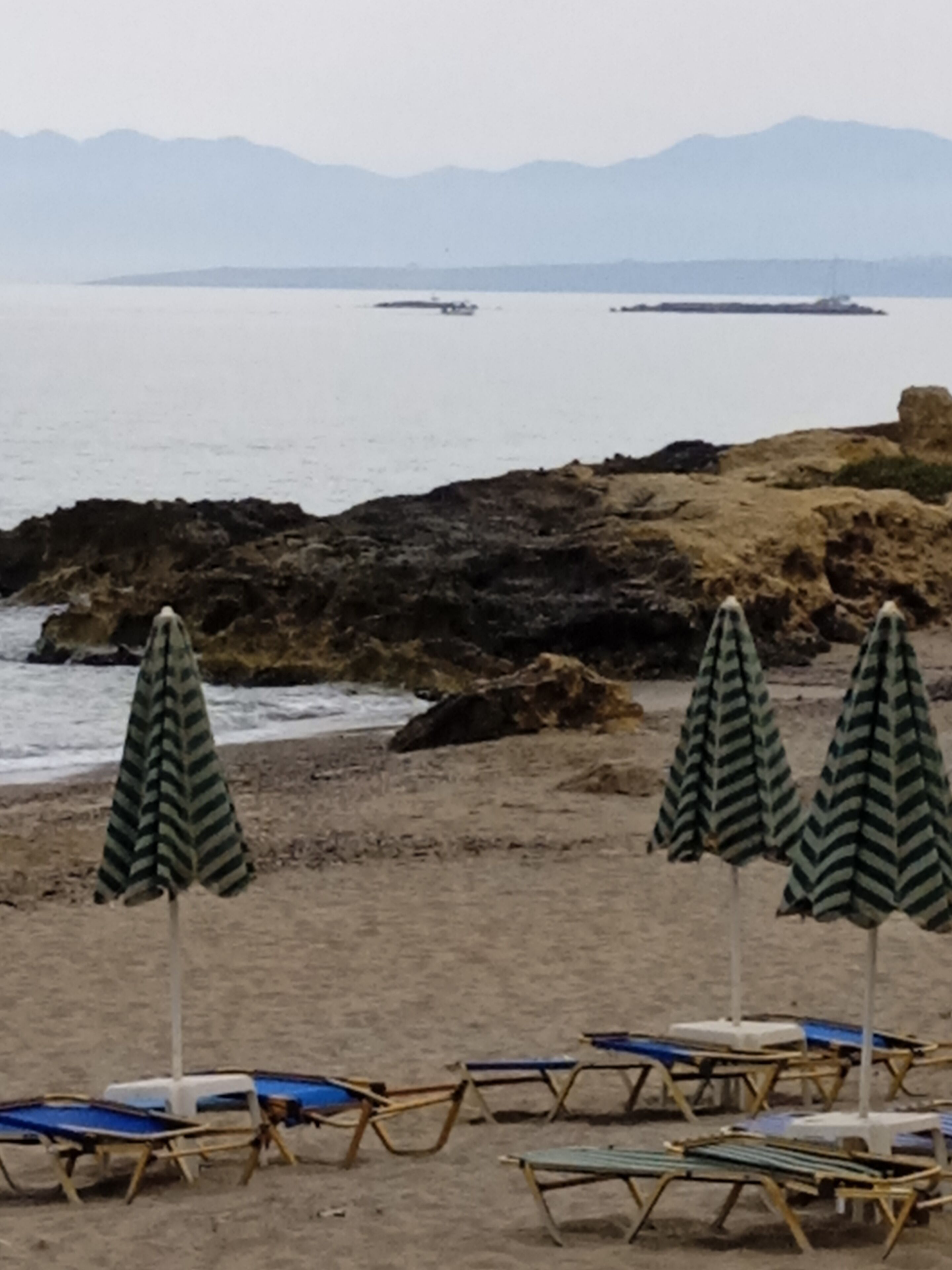 A beach with lounge chairs and striped umbrellas, rocky coastline, and distant mountains.