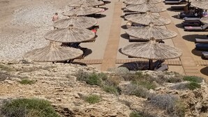 A beach with thatched umbrellas and lounge chairs, a lifeguard tower, and a view of the city in the background. - Ilona Apartments (Chania)