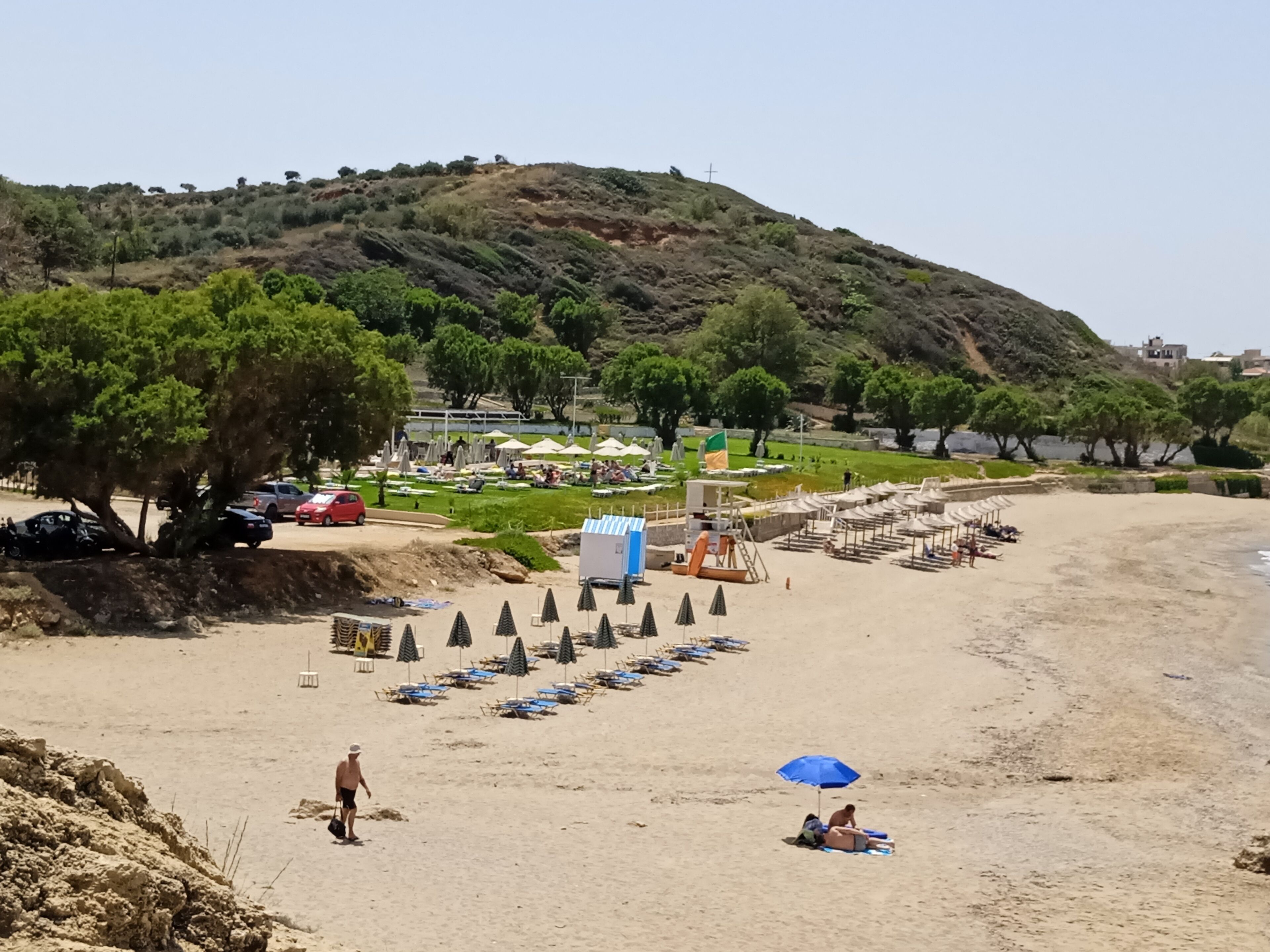 Una playa con filas de tumbonas y sombrillas, una torre de salvavidas y una colina con vegetación dispersa al fondo.
