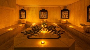 Ett spa-rum med ett stort badkar i sten som värms upp från mitten, tre dekorativa paneler på väggarna och ett flertal ljus som skapar en behaglig atmosfär.