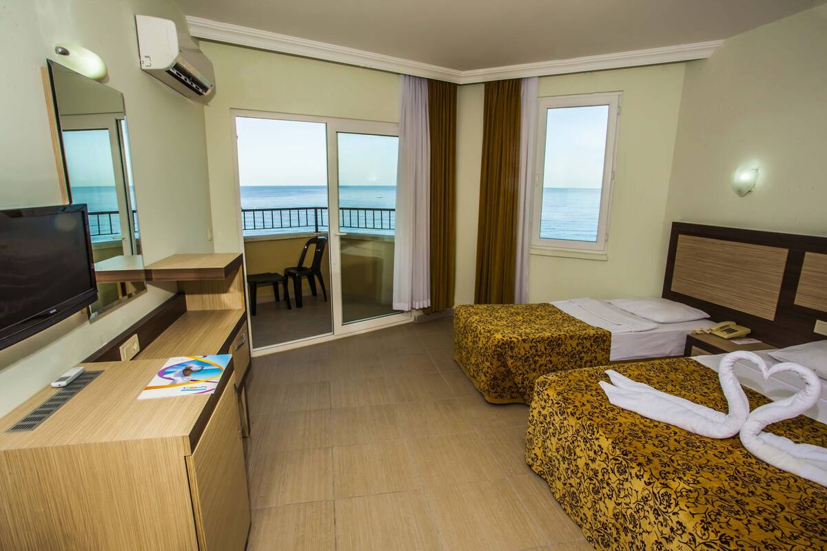 A hotel room with a balcony, a desk with a television, two beds with towels arranged in swans, and a view of the sea.