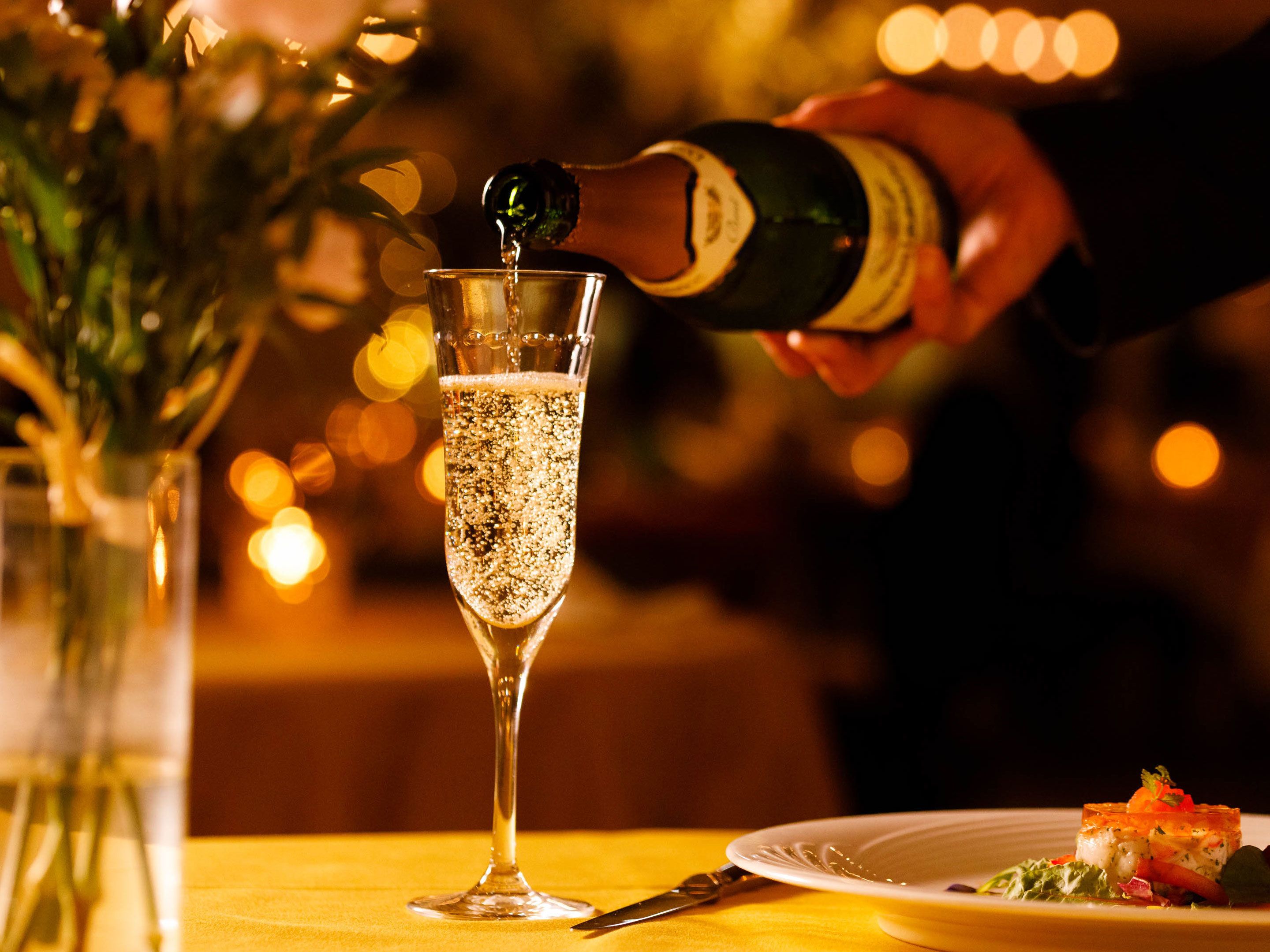 A champagne glass being filled, a plate of food, and a vase with flowers on a table.