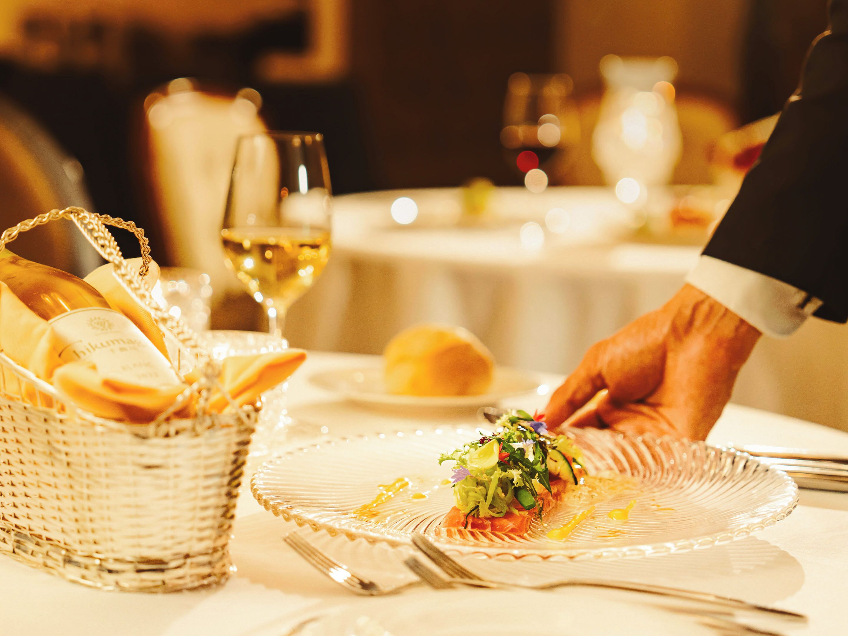 Una mesa de comedor con una cesta de pan, un plato de ensalada y una copa de vino blanco.