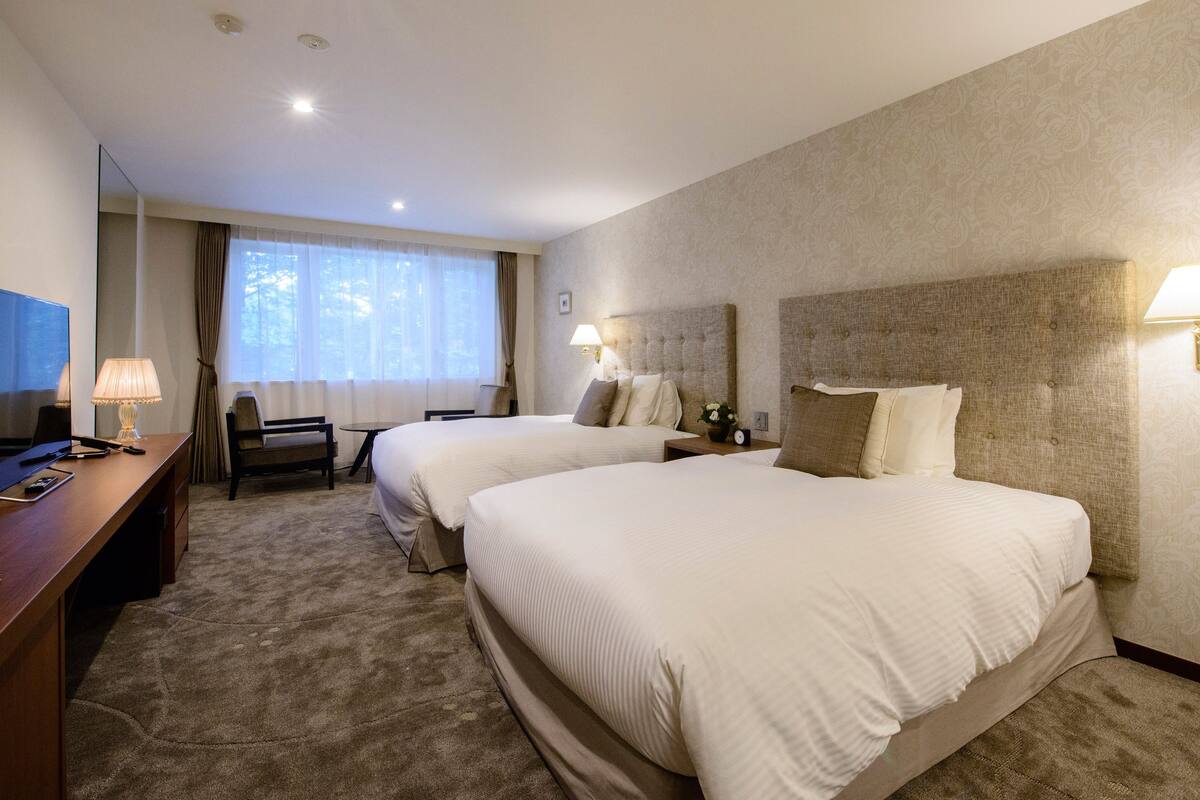 A hotel room with two beds, a desk, a chair, a TV, and a window with curtains.