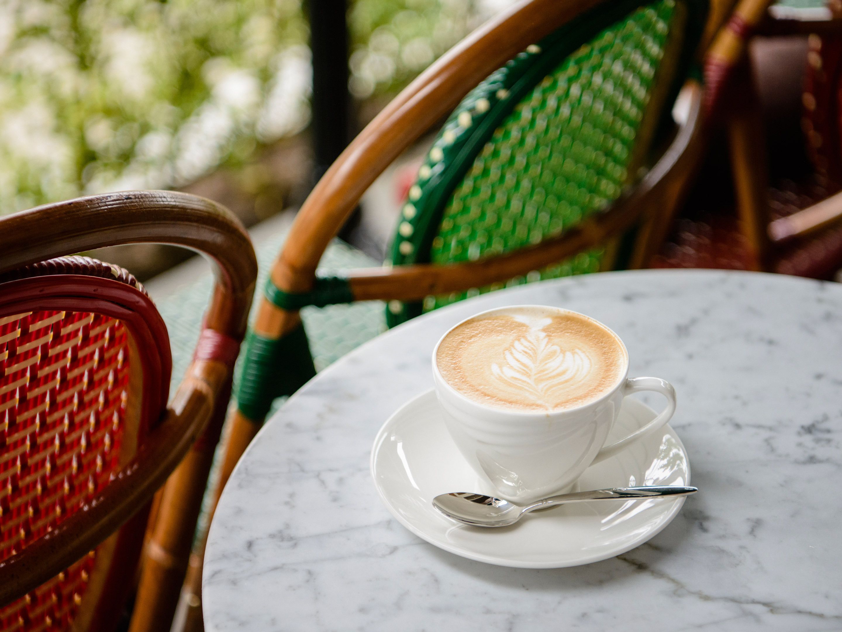 A white cup of coffee with latte art on a white saucer, accompanied by a spoon, on a marble table.