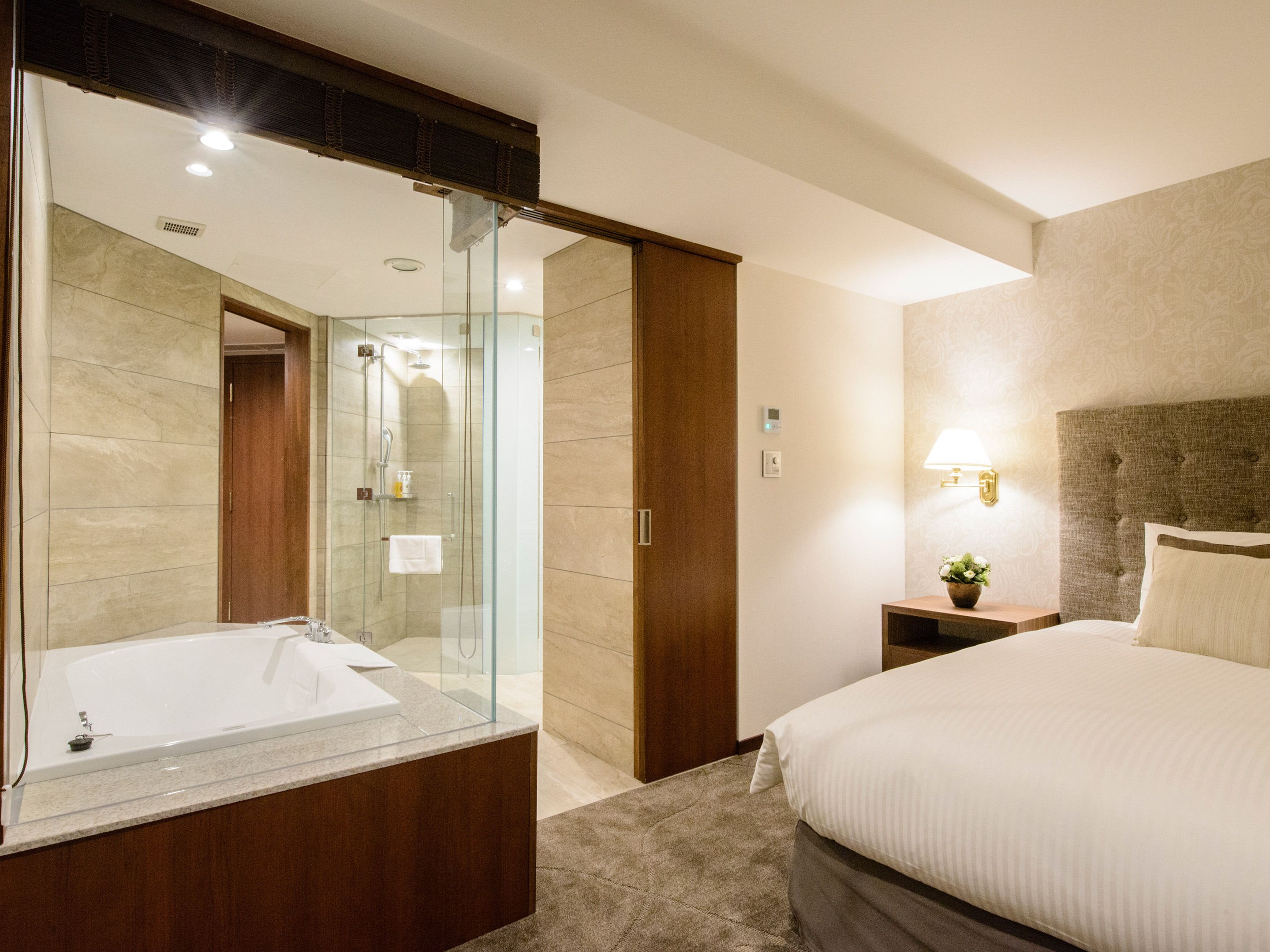 A hotel room with a large mirror, a bathtub, a bed, a bedside table with a lamp, and a small plant.