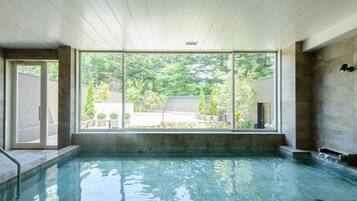 A modern indoor swimming pool with clear glass walls, a tiled floor, and a view of a garden and trees outside.