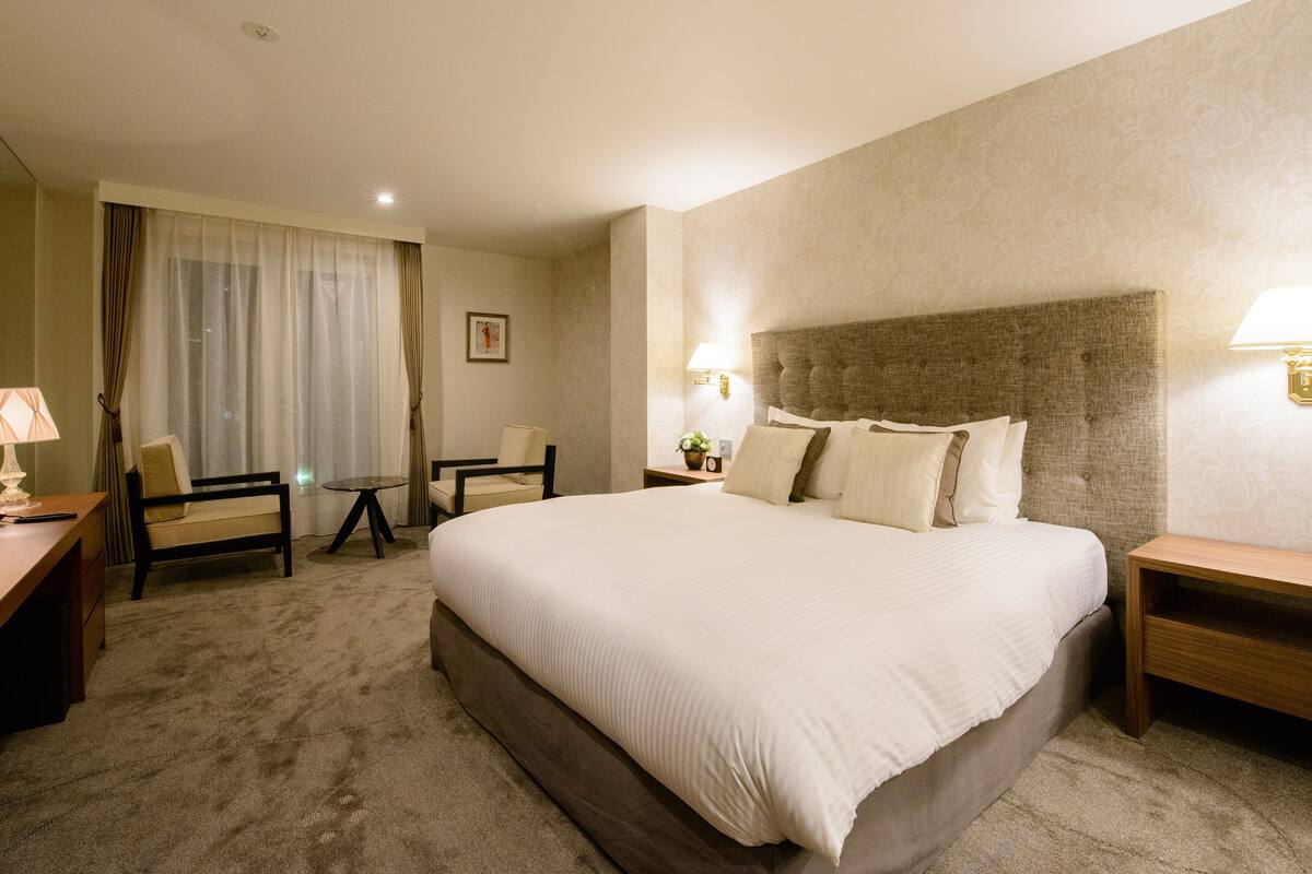 A hotel room with a large bed, two bedside tables, a desk with a chair, a small table, and a chair.