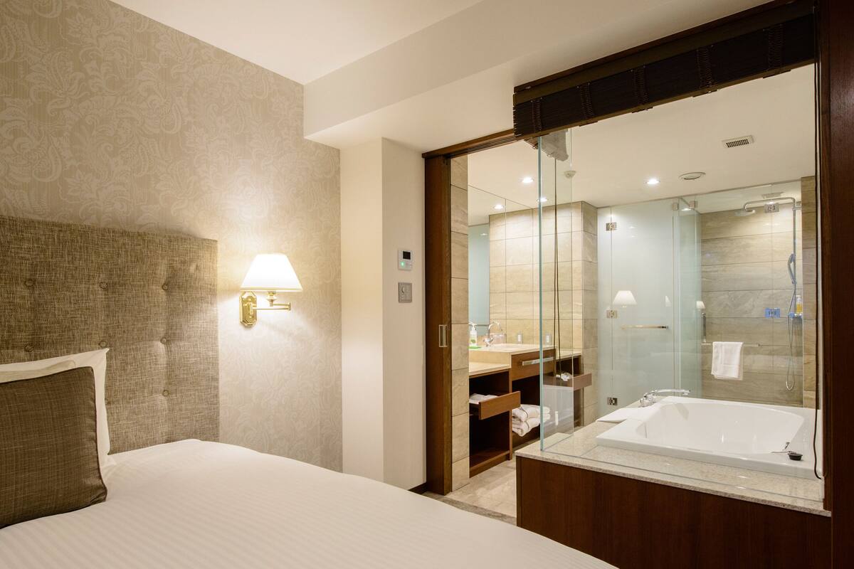 A hotel room with a large bed, a glass-enclosed shower area, and a wooden wardrobe.