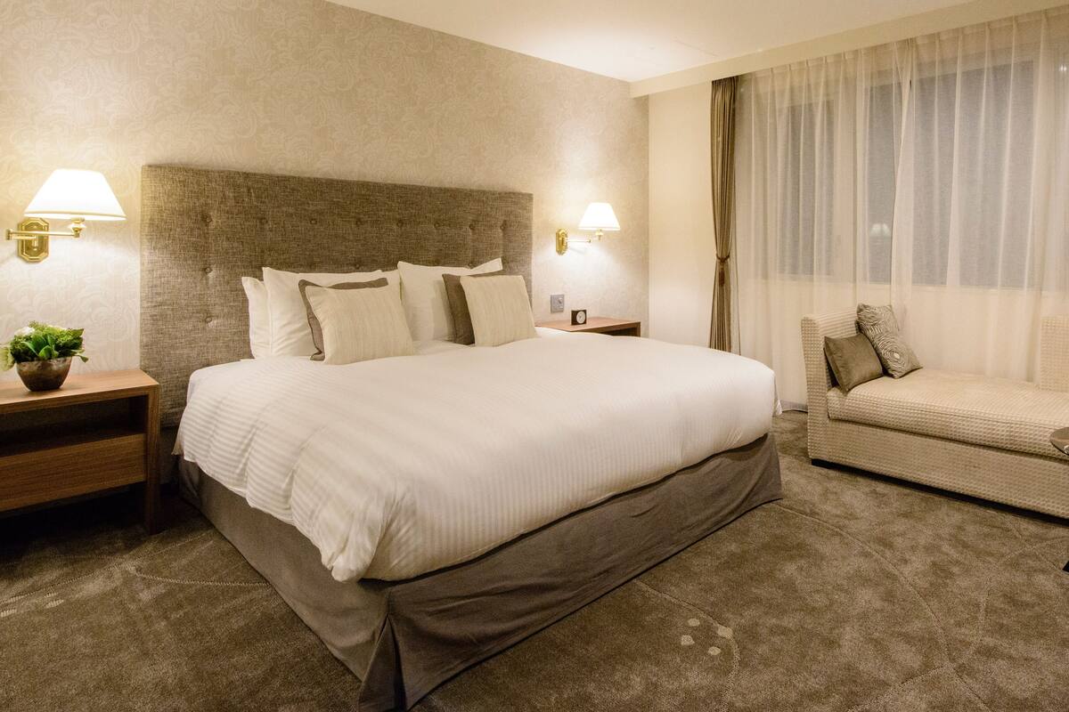 A hotel room with a large bed, a sofa, a nightstand with a lamp, and a wall-mounted lamp.
