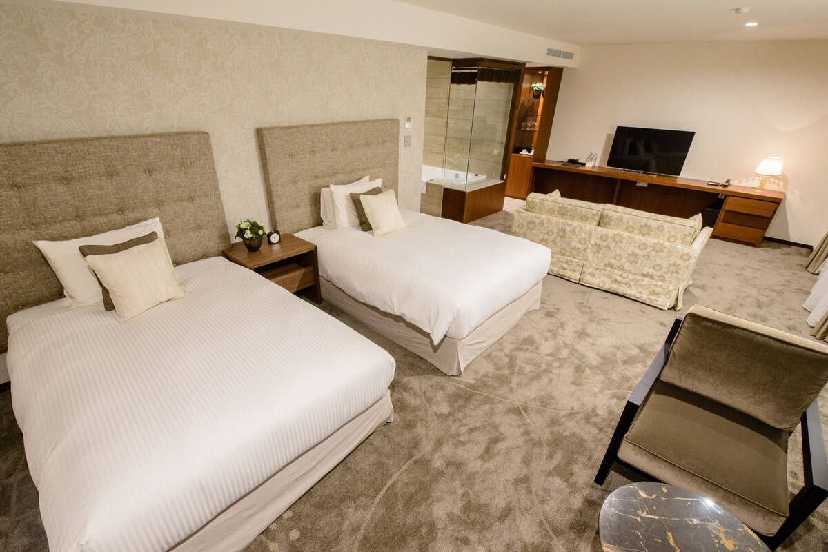 A hotel room with two beds, a sofa, a TV, a bathroom, and a marble table.
