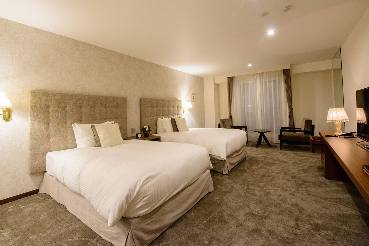 A hotel room with two beds, a desk, a chair, a TV, and a large window with curtains.