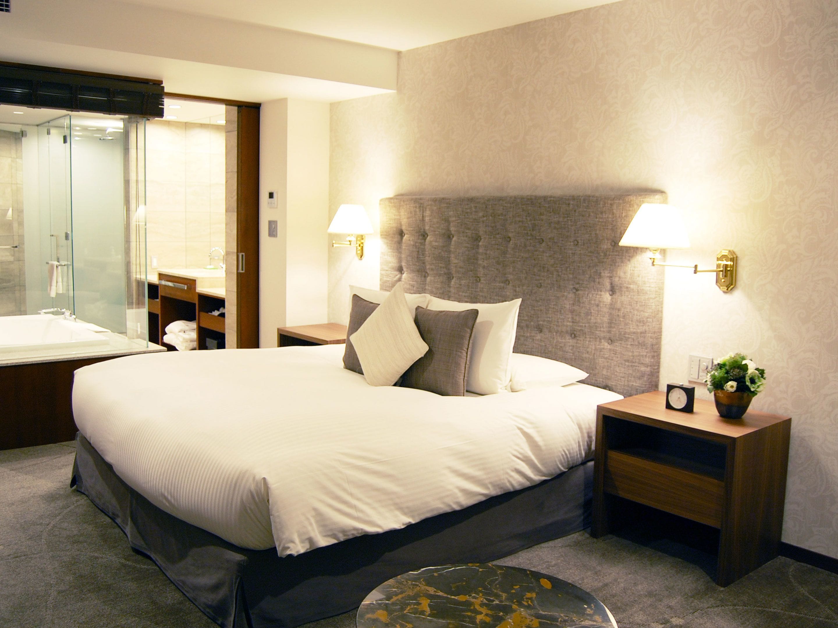 A hotel room with a large bed, bedside tables, a nightstand, a mirror, a bathroom with a glass shower enclosure, and a small desk with a chair.