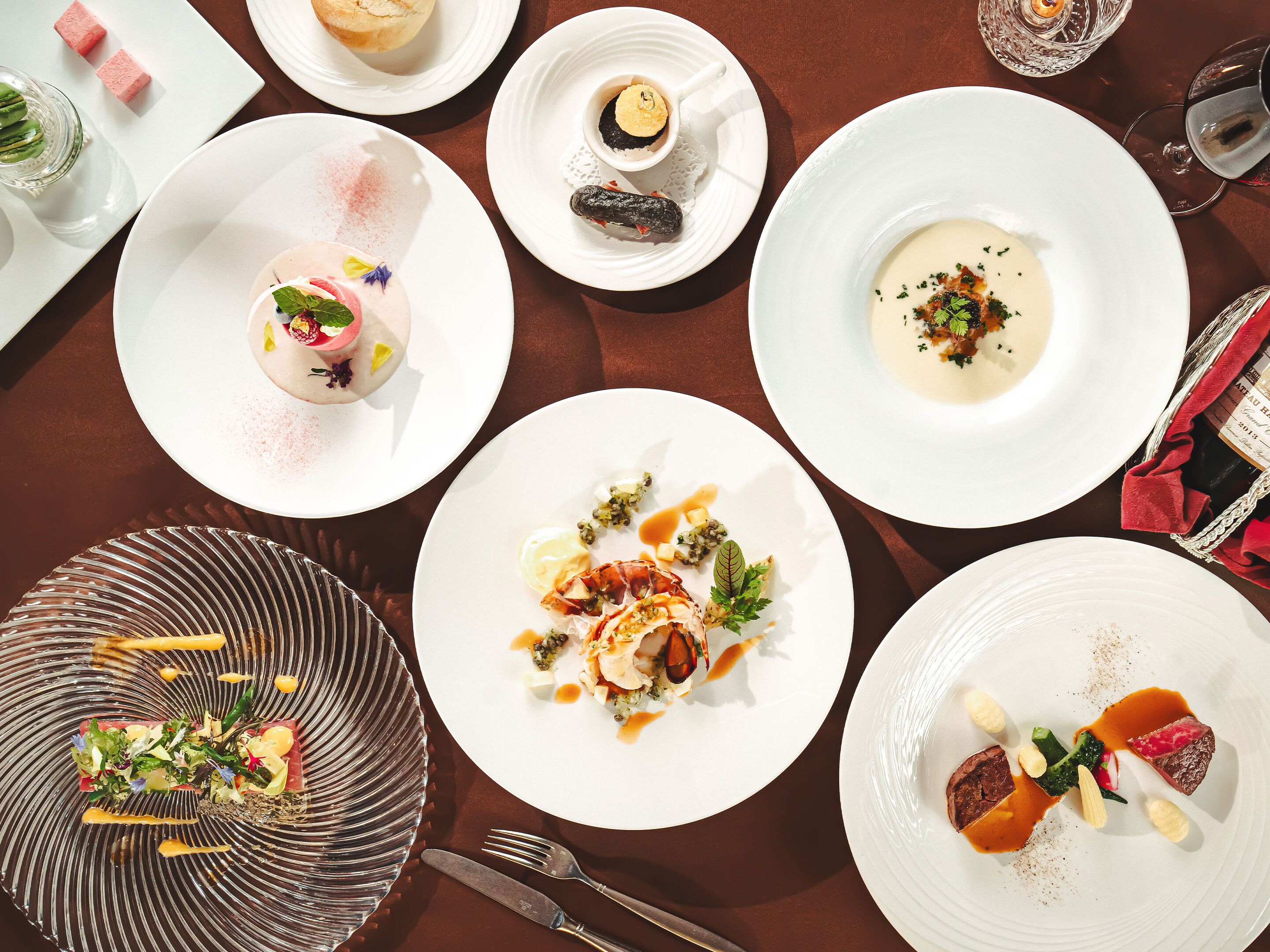 Ein gedeckter Tisch mit verschiedenen Gerichten, darunter ein Dessert mit rosa Zuckerstreuseln, eine Teller mit cremiger Suppe, garniert mit Kräutern, und ein Teller mit einem Steak und Gemüse.