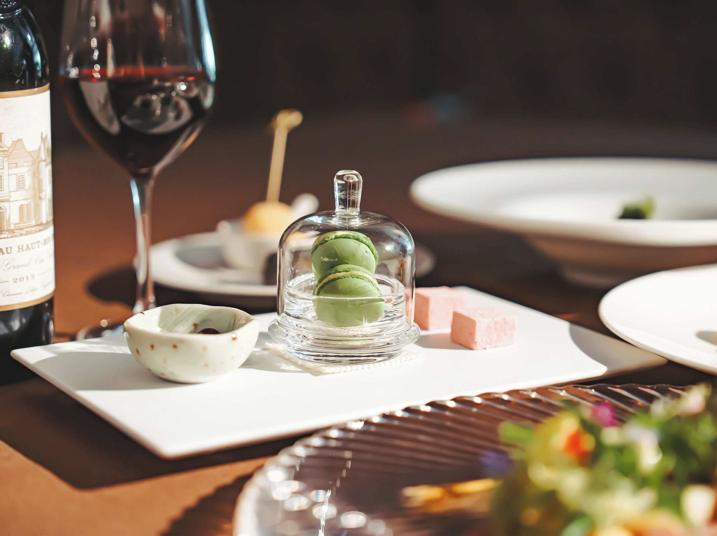A dining table with a wine bottle, a glass of red wine, a dessert with macarons, and a small bowl of sauce.