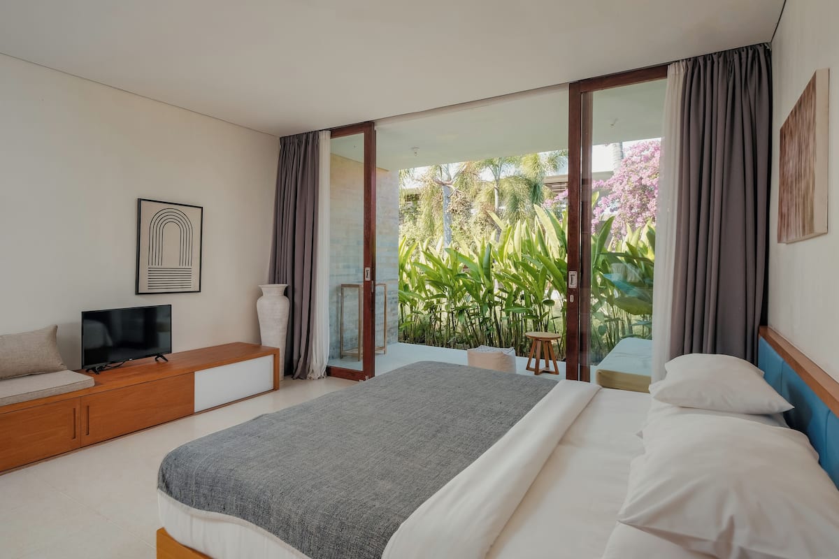 A modern hotel room with a large bed, a TV on a wooden console, a balcony with plants, and a sliding glass door.