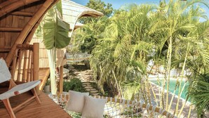 A wooden deck with a hammock and a chair, surrounded by lush palm trees and a view of a clear blue pool. - Origin Lombok (Kuta)
