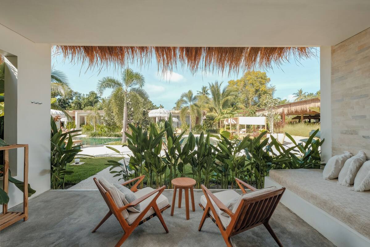 A modern outdoor seating area with a pool view, featuring wooden furniture and lush tropical plants.