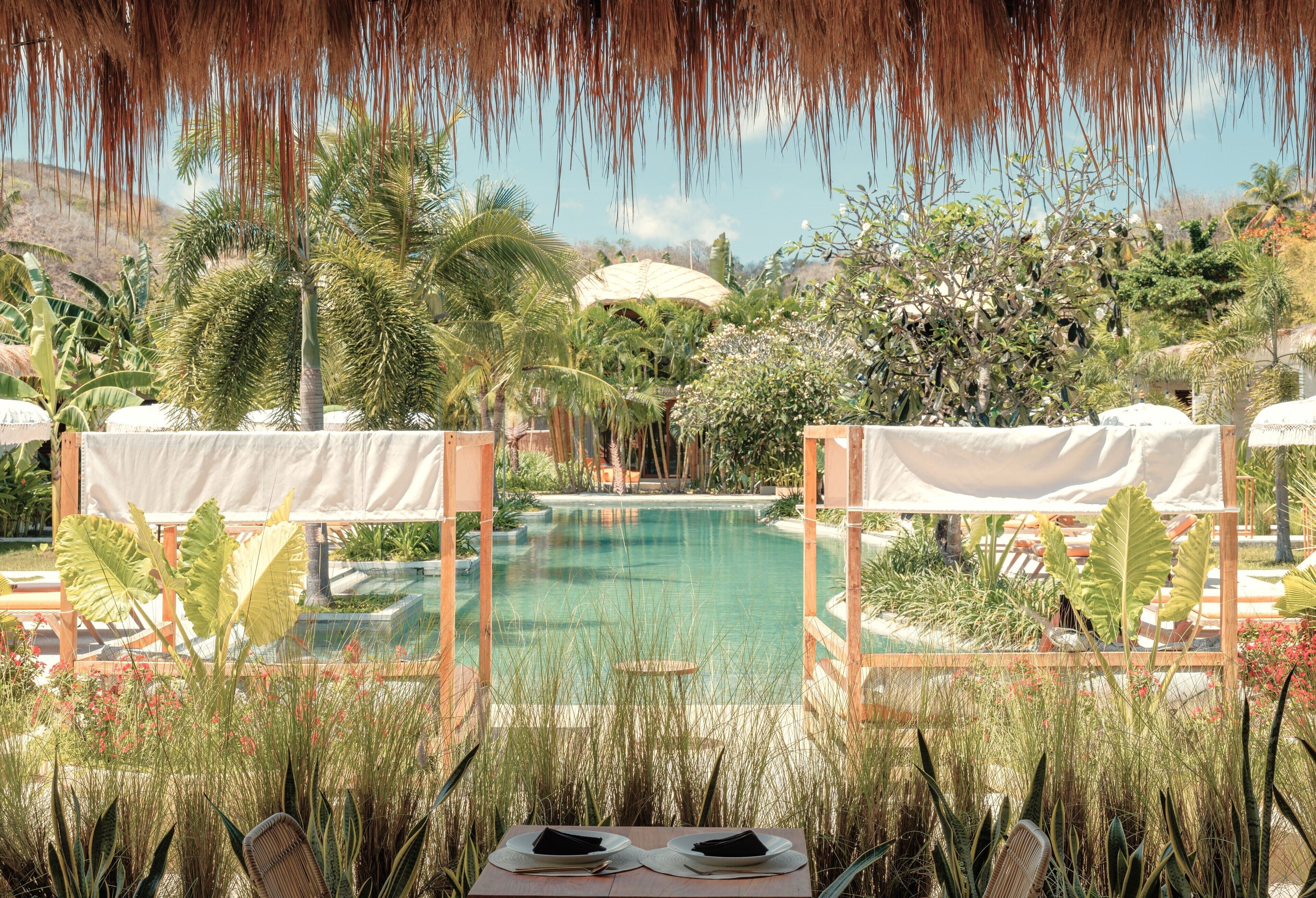 A poolside area with a clear view of the pool, surrounded by lush greenery and palm trees. There are two covered seating areas with umbrellas.