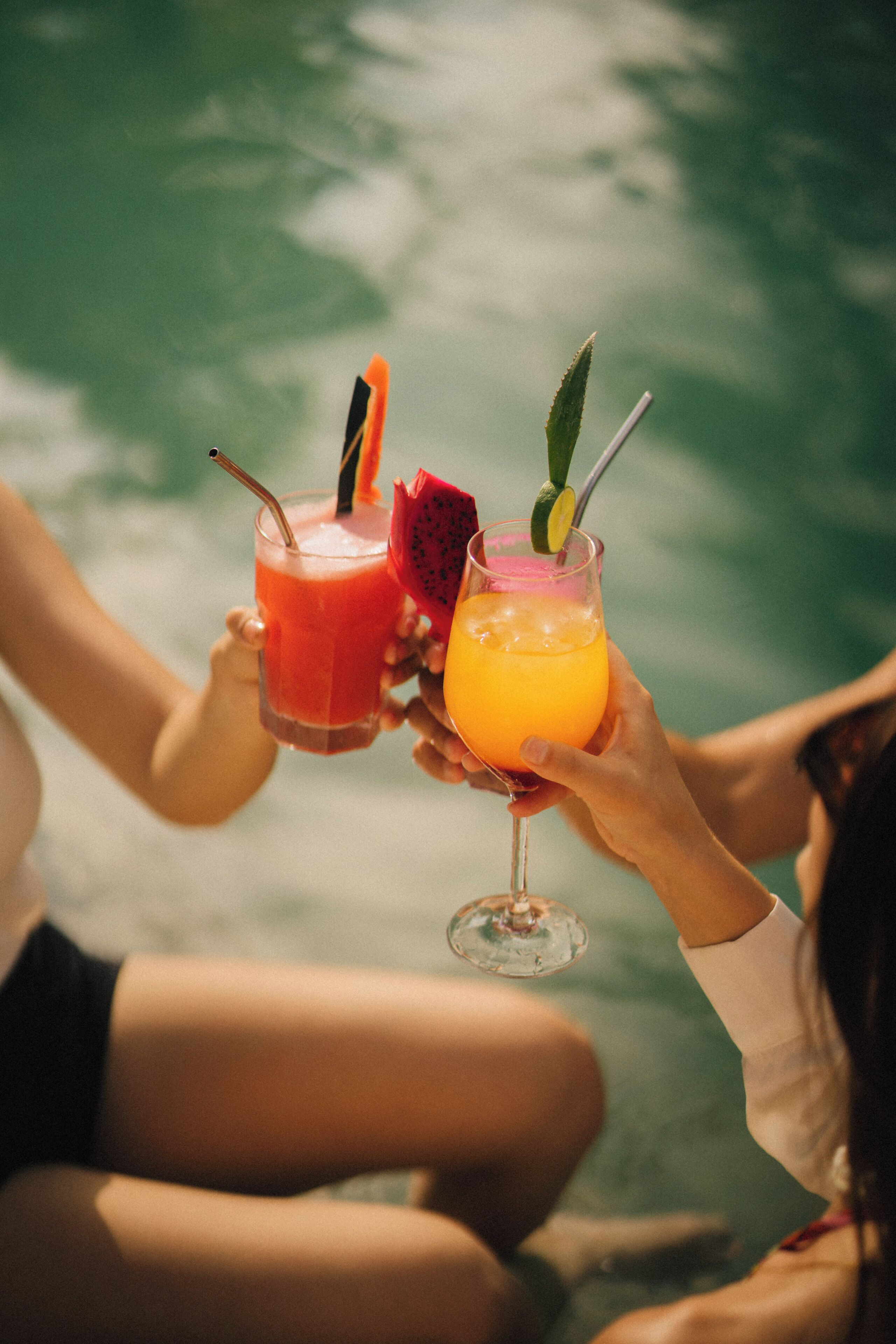 To personer holder drinks med sugerør, hvoraf den ene er pyntet med frugt, ved en swimmingpool.