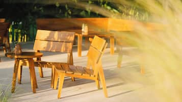 A wooden outdoor seating area with a round table and chairs, surrounded by greenery.