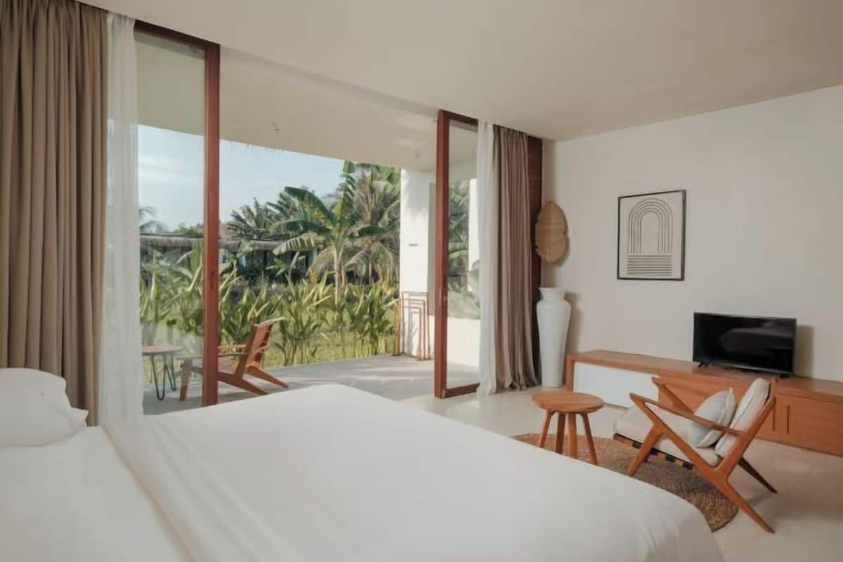 A bedroom with a large bed, a TV, a desk, and a balcony with a view of palm trees.