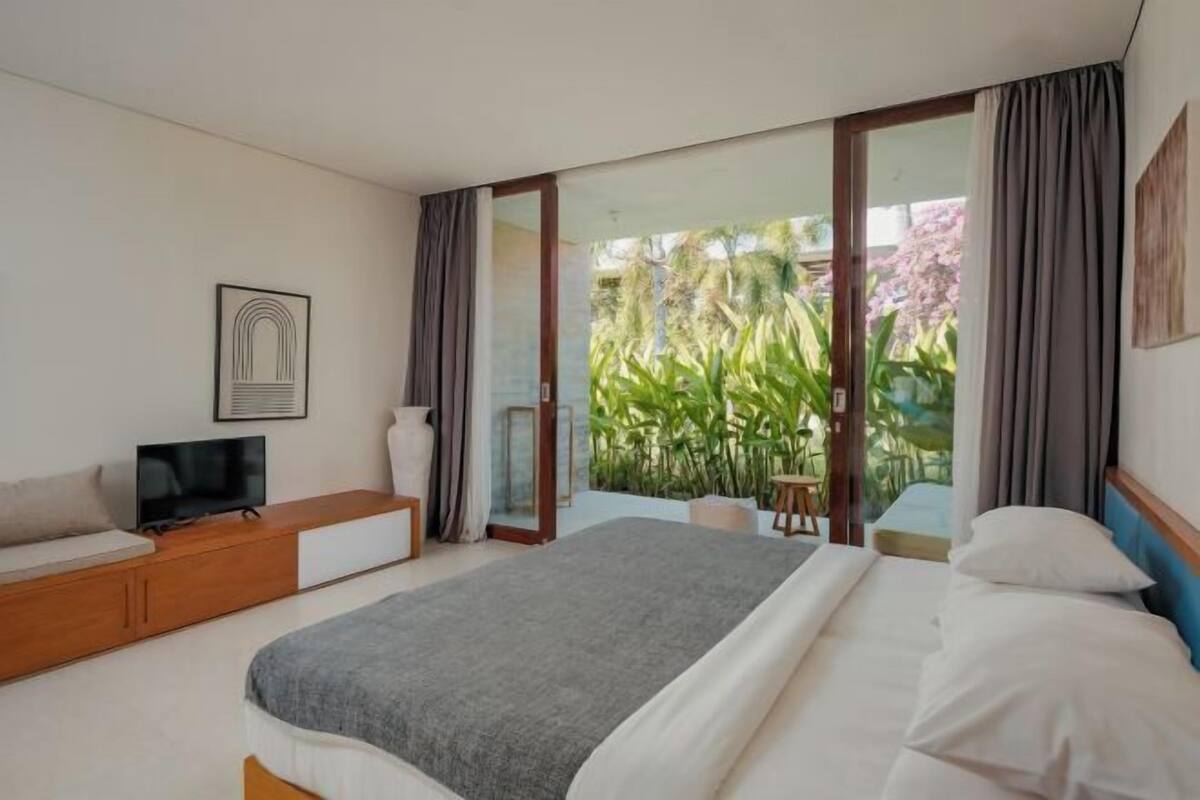 A bedroom with a bed, a TV on a wooden console, a large window with curtains, and a view of greenery outside.