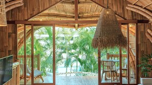A room with a large glass door leading to a tropical garden, wooden ceiling, and a hanging light fixture. - Origin Lombok (Kuta)